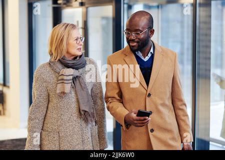 Homme d'affaires africain au manteau en utilisant un téléphone portable et en parlant à son collègue pendant qu'ils entrent dans le bâtiment de bureau Banque D'Images