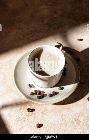 Une tasse de cappuccino ou de latte aux grains de café épars, gros plan dans une lumière dure avec des ombres sur fond beige. Copier l'espace. Orientation verticale Banque D'Images