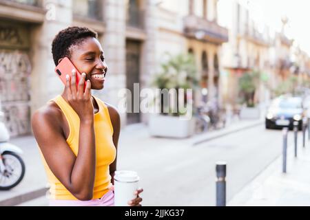 Belle femme afro-américaine debout dans la rue et souriante en parlant par téléphone. Elle porte une tenue décontractée d'été et une coupe de cheveux tendance Banque D'Images