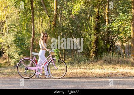 Vue latérale d'une jeune femme sur des lunettes de soleil et tenue d'été portant un vélo sur le côté à travers le parc naturel Banque D'Images
