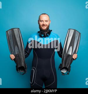 Beau barbu homme en costume de plongeur souriant et tenant les flippers sur fond bleu Banque D'Images