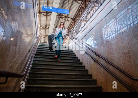 Une jeune femme descend les escaliers de la gare avec un sac à roulettes - une femme d'affaires descend les marches du terminal avec ses bagages Banque D'Images