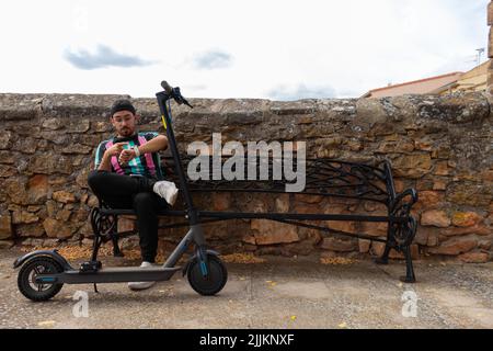 Un jeune homme caucasien assis sur un banc de métal et un mur de pierre à côté de son scooter électrique regardant sa montre Banque D'Images