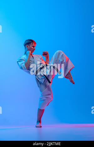 Jeune homme, chasseur de karaté dans un kimono blanc en action, mouvement isolé sur fond bleu au néon. Sport, action, sports de combat, concept publicitaire Banque D'Images