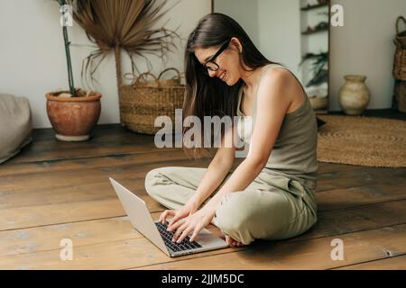 Jeune fille utilise un ordinateur portable pour l'étude en ligne assis sur un plancher en bois. Banque D'Images