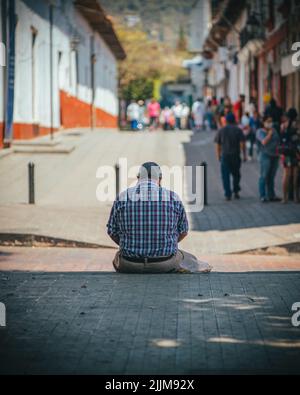 Une photo verticale d'un homme âgé dans une chemise à carreaux assis sur un trottoir. Vue de l'arrière Banque D'Images