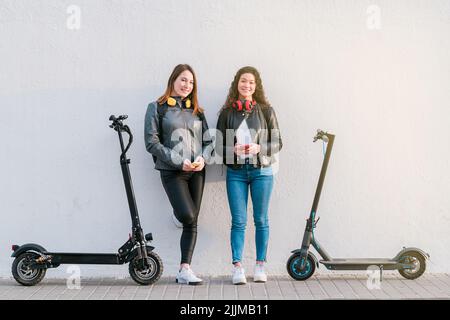 Deux amies multiethniques sur des scooters électriques utilisant un smartphone à l'extérieur. Banque D'Images