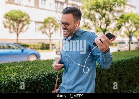 Des écouteurs professionnels qui crient et chantent tout en faisant des gestes. Les jeunes hommes qui se transportent apprécient la musique sur leur smartphone. Il marche sur le côté Banque D'Images