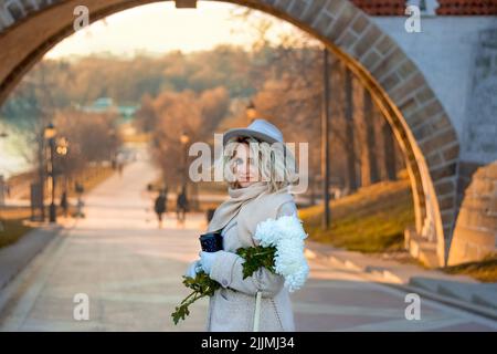 Une femme blonde dans un manteau, un chapeau et des gants avec un bouquet de chrysanthèmes blancs et une tasse de café sur le fond d'une arche et d'une landsca d'automne Banque D'Images
