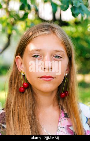 Refocalisation belle blonde jeune fille avec cerise douce sur l'oreille. Belle adolescente souriante sur fond vert. Boucles d'oreilles pour enfants. Portrai d'été Banque D'Images