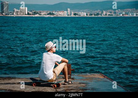 Un jeune caucasien en lunettes de soleil assis sur un skateboard sur un quai en béton qui regarde une mer Banque D'Images