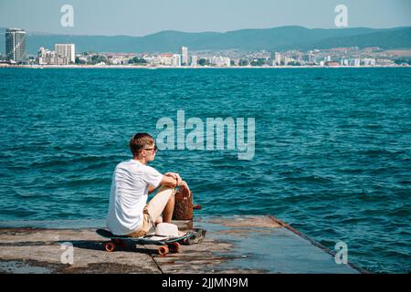 Un jeune caucasien en lunettes de soleil assis sur un skateboard sur un quai en béton qui regarde une mer Banque D'Images