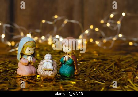 Un gros plan de petites figures colorées de la scène Nativité sur foin avec une série de lumières et un mur en bois brun dans le fond flou Banque D'Images