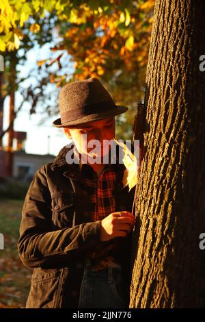 Portrait flou artistique de la jeune femme caucasienne souriante dans un parc d'automne orange coloré. Femme brillante et élégante en chapeau et chemise près du tronc d'arbre, érable jaune Banque D'Images