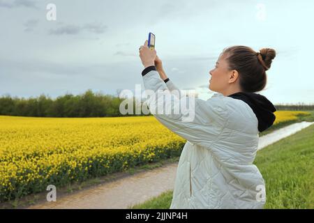 petite et belle fille tourne une vidéo d'un champ de colza jaune sur son téléphone. La fille se repose dans la nature et admirent le paysage envoûtant Banque D'Images