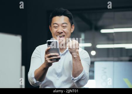 Une pause au travail. Un jeune asiatique tient un téléphone dans ses mains, joue au téléphone au bureau au travail, se réjouit, montre avec sa main oui, se réjouit de la victoire, a gagné un prix, repose, ne fonctionne pas. Banque D'Images