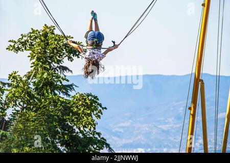 Petite fille avec jean short et cheveux longs à l'envers sur balançoire élastique rebondissant surplombant la montagne et la vallée Banque D'Images