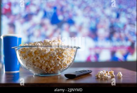 Sur fond de stade avec des spectateurs sur l'écran de télévision, un grand bol de pop-corn en verre et une télécommande de télévision. Détendez-vous devant Banque D'Images