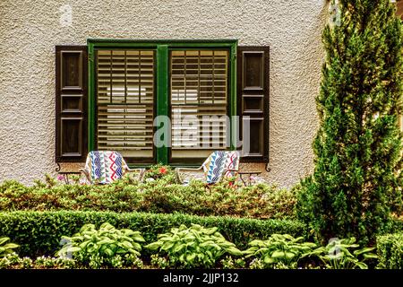 Salon extérieur avec salon, mobilier de jardin devant une fenêtre avec volets et derrière des plantes de jardin paysagées, y compris des haies et des hostas Banque D'Images