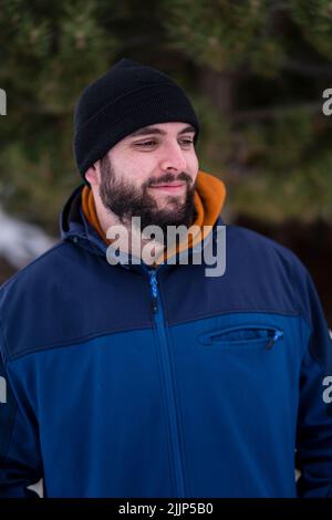 Une photo verticale d'un jeune homme caucasien dans une veste d'hiver bleue debout dans un parc et souriant Banque D'Images