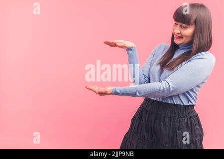 Jeune femme brune caucasienne avec des langes portant un col de cygne bleu avec ses mains et regardant à gauche. Prise de vue en studio sur fond rose. . Photo de haute qualité Banque D'Images