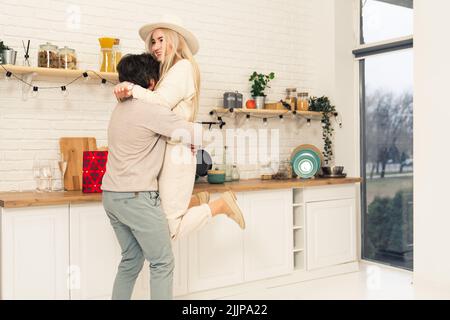 Brunette homme ramassant la femme blonde souriante du sol et l'embrassant dans la cuisine. Couple caucasien. Prise de vue en intérieur. Photo de haute qualité Banque D'Images