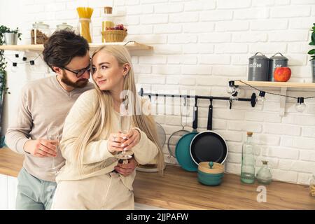 Un couple blanc, un homme brunette et une femme blonde dans la cuisine se tenant en regardant les yeux de l'autre en souriant et en buvant du champagne. Photo Indooe. . Photo de haute qualité Banque D'Images
