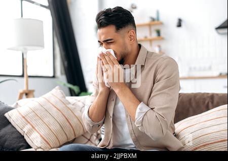 Le concept de la maladie, l'écoulement nasal. Un homme arabe ou indien malsain et triste est assis à la maison dans la salle de séjour sur le canapé, se sent mal, souffre d'un nez qui coule, des éternuements, utilise des serviettes, a besoin de traitement Banque D'Images