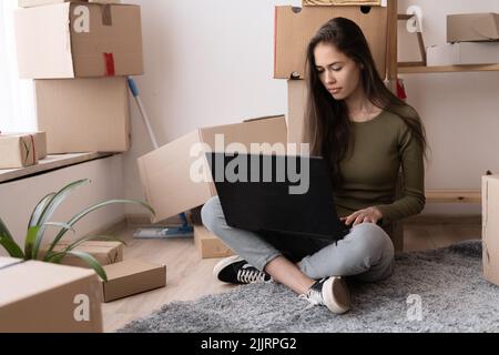 fille heureuse avec des boîtes se déplace à nouvel appartement, se déplaçant à une nouvelle maison, logement de location, jeune femme espagnole utilisant ordinateur portable pour rechercher et commander un Banque D'Images