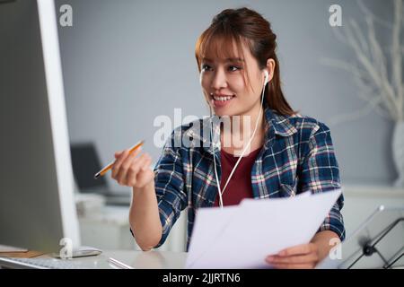 Manager asiatique joyeux écoutant de la musique dans un casque tout en étant assis devant l'ordinateur et en travaillant sur le projet de démarrage, portrait de taille haute Banque D'Images