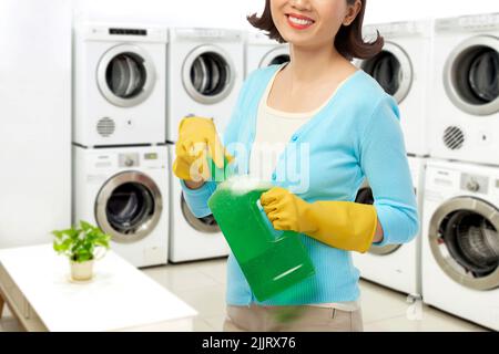 Jeune femme souriante portant des gants en caoutchouc ouvrant une bouteille de détergent liquide tout en se tenant dans une grande buanderie libre-service Banque D'Images