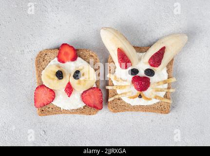 Deux toasts doux pour les enfants en forme de poussin et de lapin avec fraises, banane, fromage à la crème et flocons de noix de coco sur fond gris, vue du dessus Banque D'Images