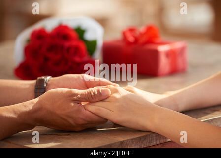 Gros plan d'un homme méconnaissable tenant les mains de sa petite amie, bouquet de roses rouges et boîte cadeau rouge couchée sur une table en bois Banque D'Images