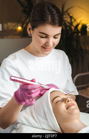 La fille subit une procédure cosmétique avec un support d'appareil par esthéticienne au salon de beauté. Soin du visage des filles. Salon de beauté/beauté. Banque D'Images