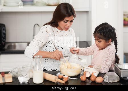 Vue de profil de jolie petite fille avec deux tresses ajoutant de la farine dans un bol tout en cuisant une tarte appétissante avec maman, intérieur de la cuisine moderne sur le fond Banque D'Images