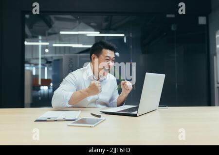Un jeune homme asiatique réussi et heureux, homme d'affaires, heureux, montre avec la main oui. A conclu un contrat. A gagné de l'argent. Assis avec un carnet à la table dans un bureau moderne Banque D'Images
