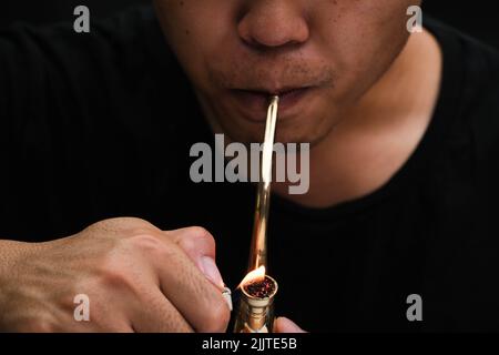 L'homme asiatique fume de la marijuana dans une pipe à la maison. Studio Shoot avec modèle simulant pot à fumer avec un tuyau dans un arrière-plan sombre. Légalisation du cannabis Banque D'Images