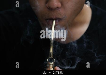 L'homme asiatique fume de la marijuana dans une pipe à la maison. Studio Shoot avec modèle simulant pot à fumer avec un tuyau dans un arrière-plan sombre. Légalisation du cannabis Banque D'Images