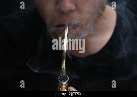 L'homme asiatique fume de la marijuana dans une pipe à la maison. Studio Shoot avec modèle simulant pot à fumer avec un tuyau dans un arrière-plan sombre. Légalisation du cannabis Banque D'Images