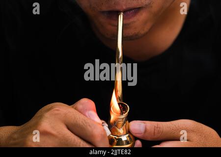 L'homme asiatique fume de la marijuana dans une pipe à la maison. Studio Shoot avec modèle simulant pot à fumer avec un tuyau dans un arrière-plan sombre. Légalisation du cannabis Banque D'Images