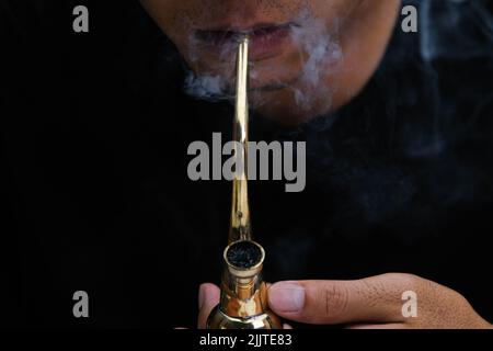 L'homme asiatique fume de la marijuana dans une pipe à la maison. Studio Shoot avec modèle simulant pot à fumer avec un tuyau dans un arrière-plan sombre. Légalisation du cannabis Banque D'Images