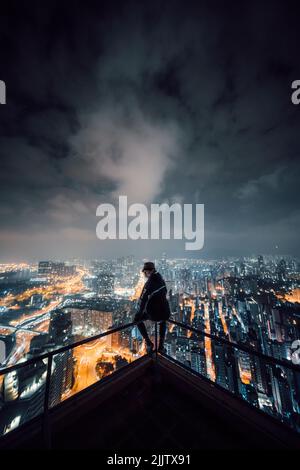 Vue arrière d'un homme asiatique assis sur un toit surplombant la ville de Hong Kong la nuit Banque D'Images
