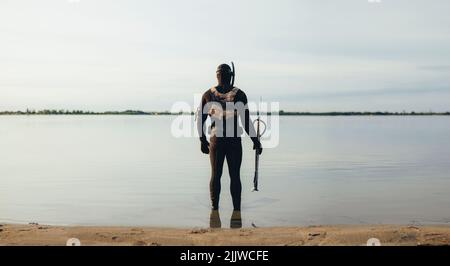 Vue arrière d'un homme debout dans l'eau de mer portant une combinaison et tenant un fer de lance. Un jeune homme aventureux se prépare à aller pêcher sous l'eau Banque D'Images