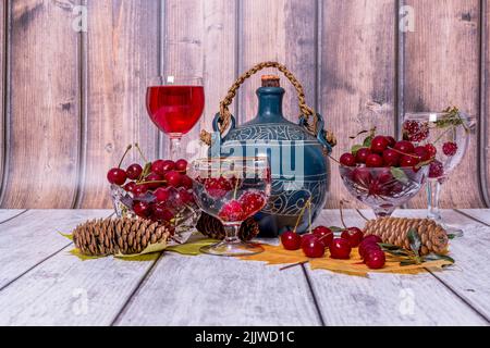 Sur la table en bois se trouvent des vases avec des cerises mûres, une carafe et un verre de vin, un verre d'eau minérale, des feuilles de coin jaunes et des cônes de sapin. Banque D'Images