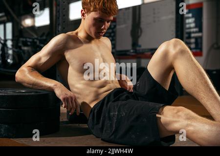Portrait du jeune homme sportif, se reposant après une séance d'entraînement dure. Assis sur le sol isolé sur fond de salle de gym Banque D'Images