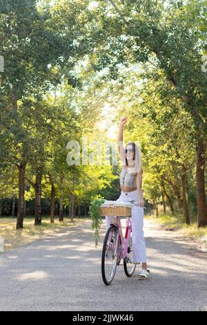 Vue verticale de la jeune femme s'amusant à monter sur un vélo rose vintage avec panier et bras montant de plante Banque D'Images
