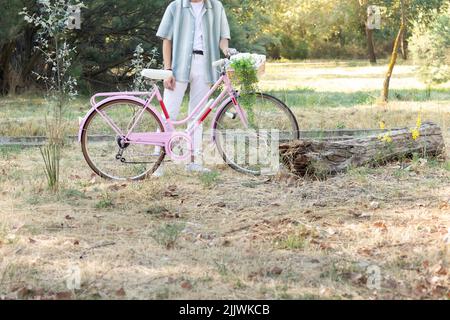 Vue latérale courte d'un jeune homme fashinable méconnaissable tenant une bicyclette rose avec un panier en osier et des plantes Banque D'Images