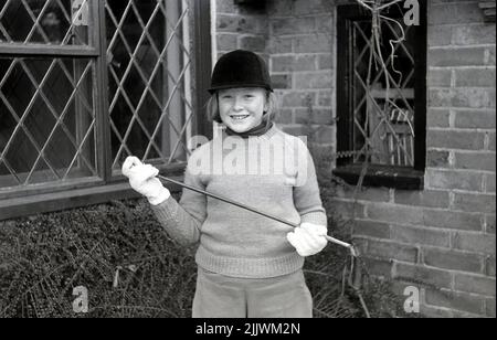 1966, historique, jeune fille heureuse debout pour une photo à l'extérieur d'une maison dans ses vêtements d'équitation, jodhpurs, gants de coton et chandail, chapeau de protection et tenir une récolte d'équitation, Angleterre, Royaume-Uni. Banque D'Images