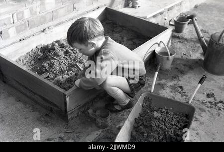 1960s, historique, à l'extérieur sur un patio, un petit garçon dans des shorts et des sandales de tartan, géotisant le sable de sa sdanpit pour mettre inot son brouette Angleterre, Royaume-Uni. Banque D'Images