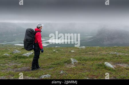 Femme randonneur dans une veste rouge avec un sac à dos lourd surplombant la vallée de la rivière Rapa dans le paysage arctique du parc national de Sarek, en Suède, sur un ciel nuageux et pluvieux Banque D'Images